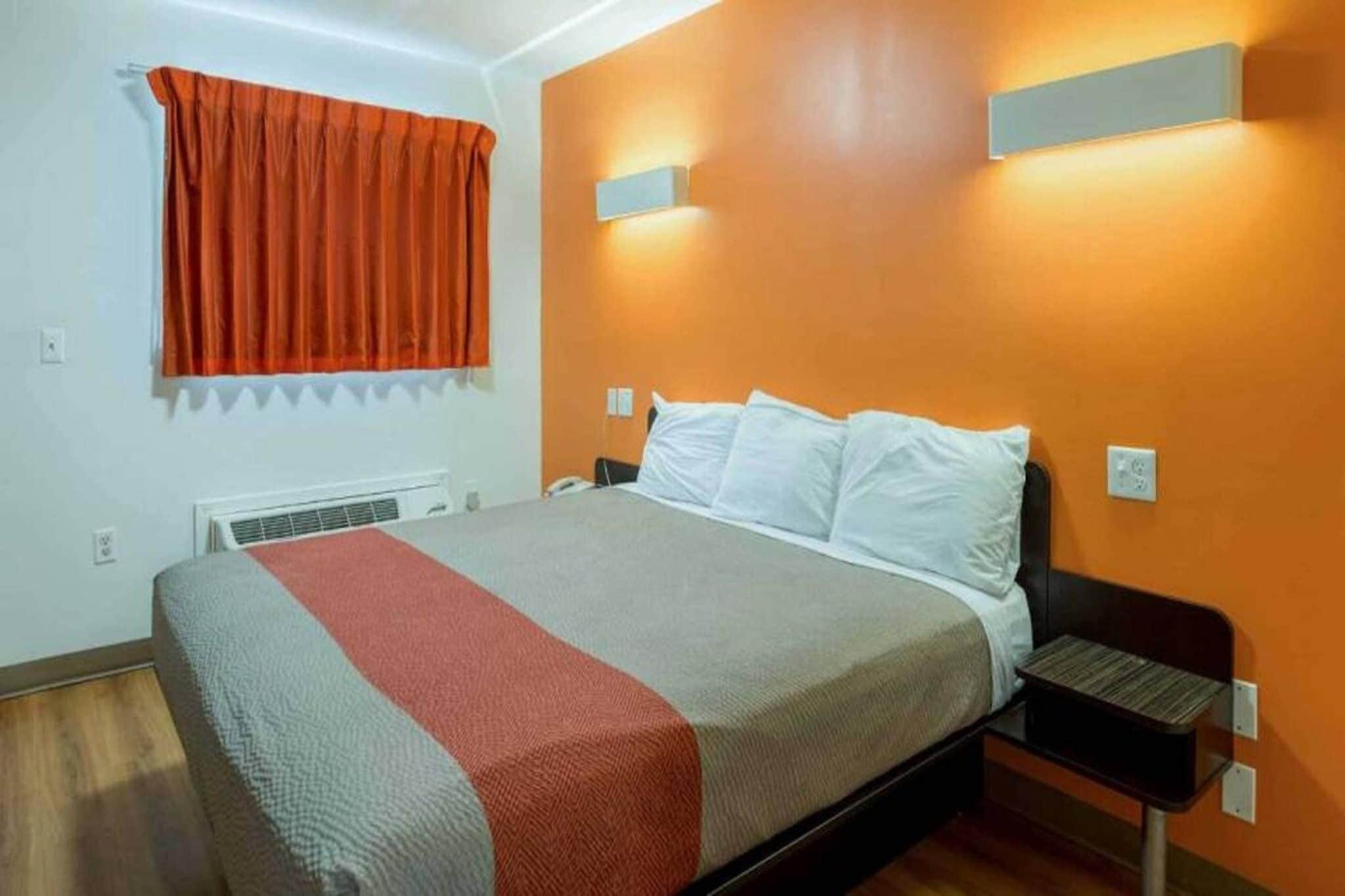 A hotel room with a bed, a nightstand, a window with curtains, and a wall-mounted light fixture.