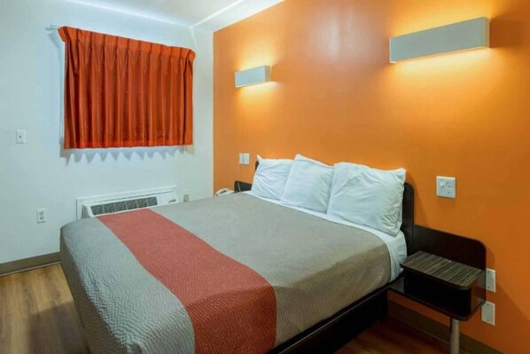 A hotel room with a bed, a nightstand, a window with curtains, and a wall-mounted light fixture.