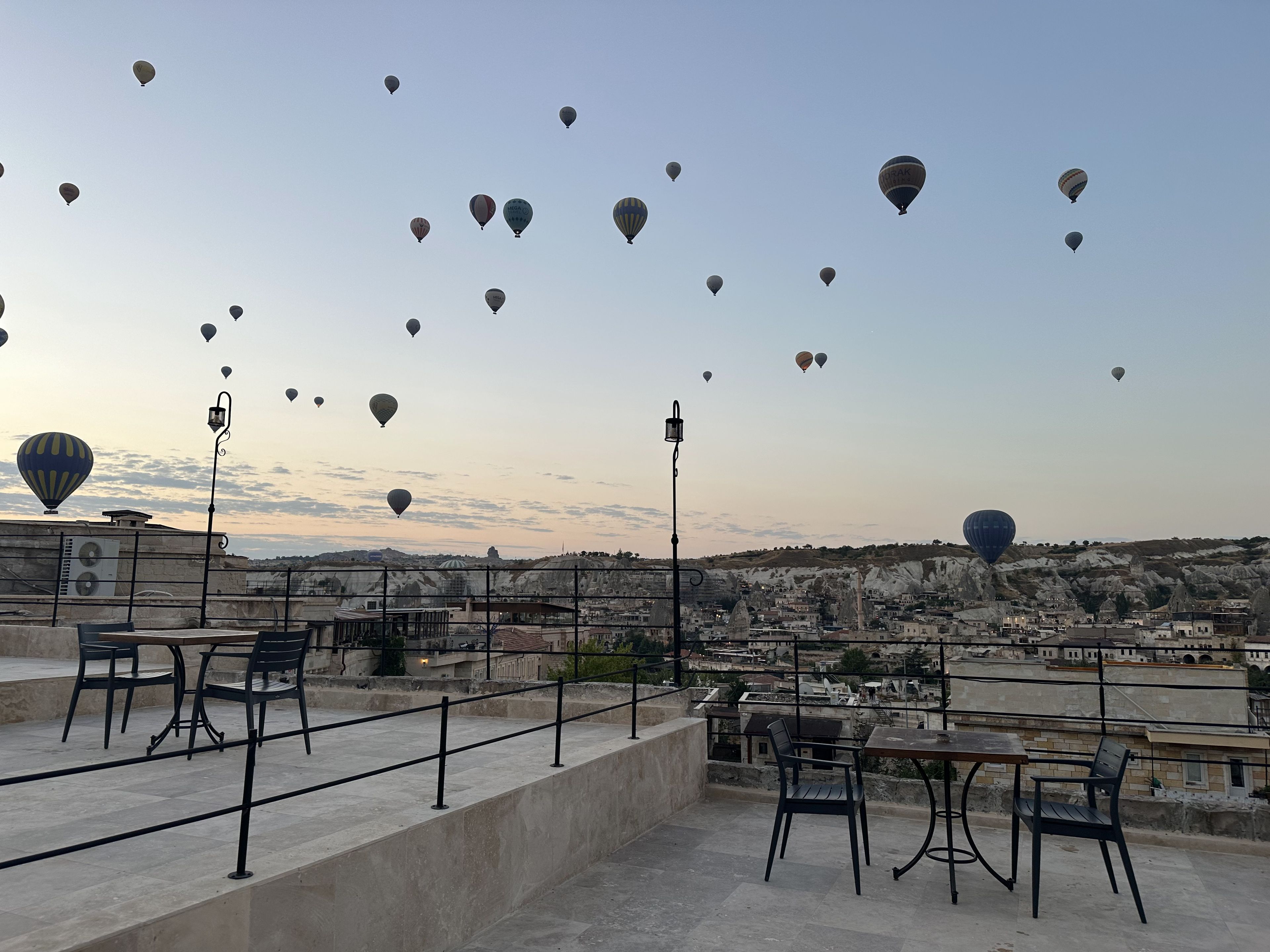 A rooftop terrace with tables and chairs, overlooking a cityscape with buildings and a mountainous backdrop.
