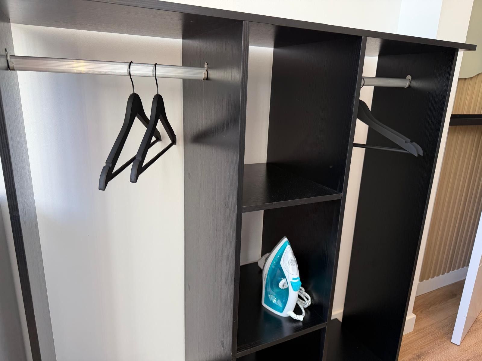 A room with a black and white shelving unit, a hanging clothes rack, and an iron on the lower shelf.