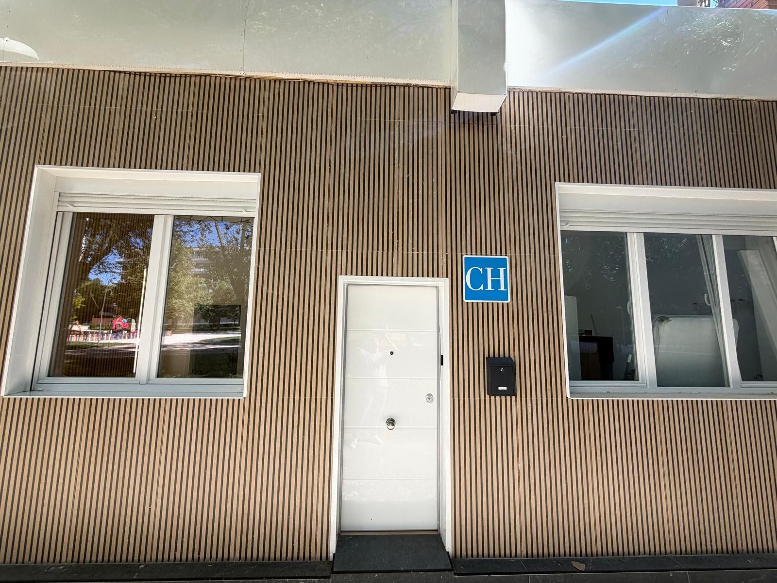Fachada de edificio con listones de madera verticales, una puerta blanca y un letrero azul con la letra 'CH'.
