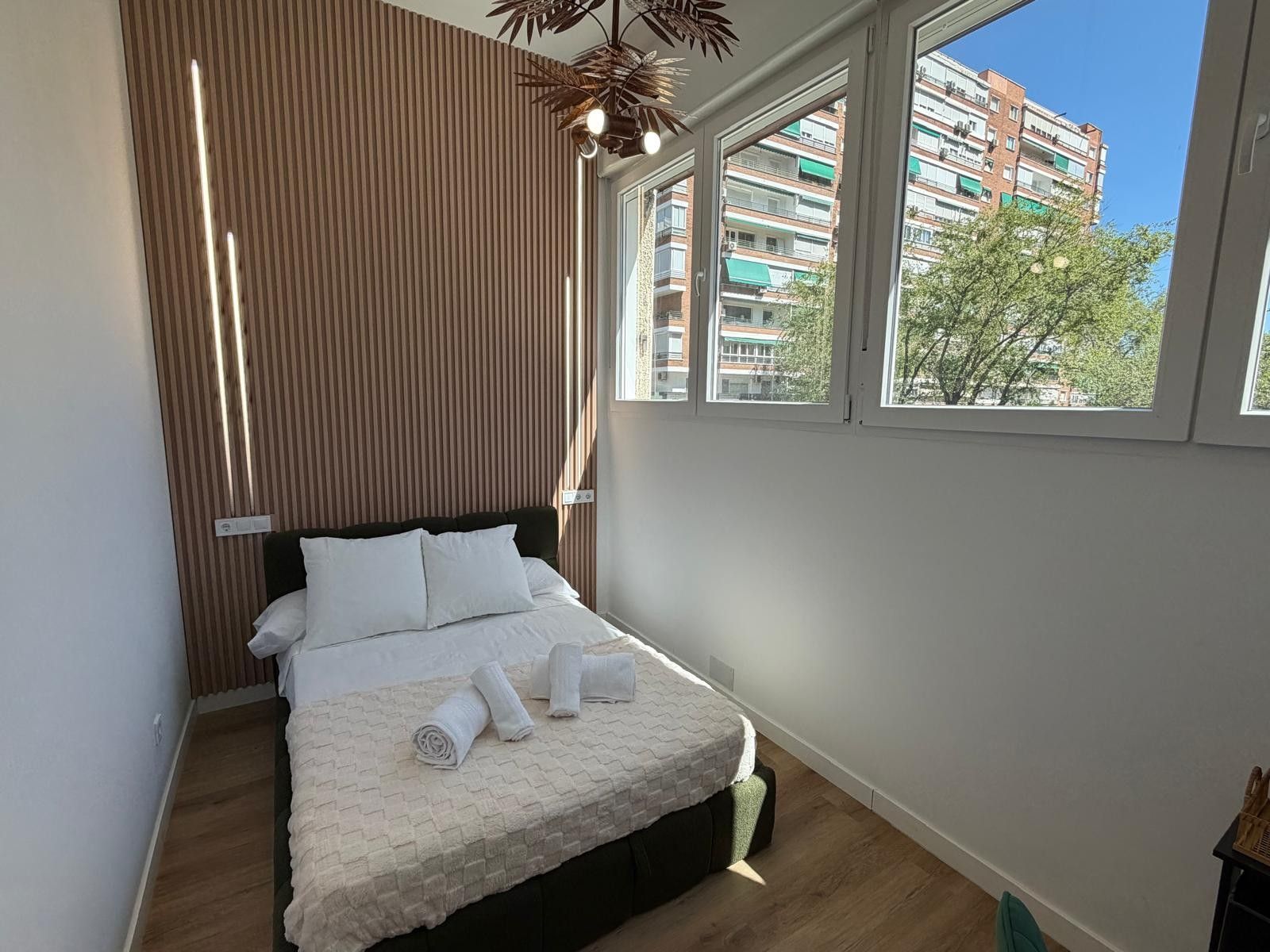 Un dormitorio con cama, pared de listones de madera, ventana y vista a edificios.