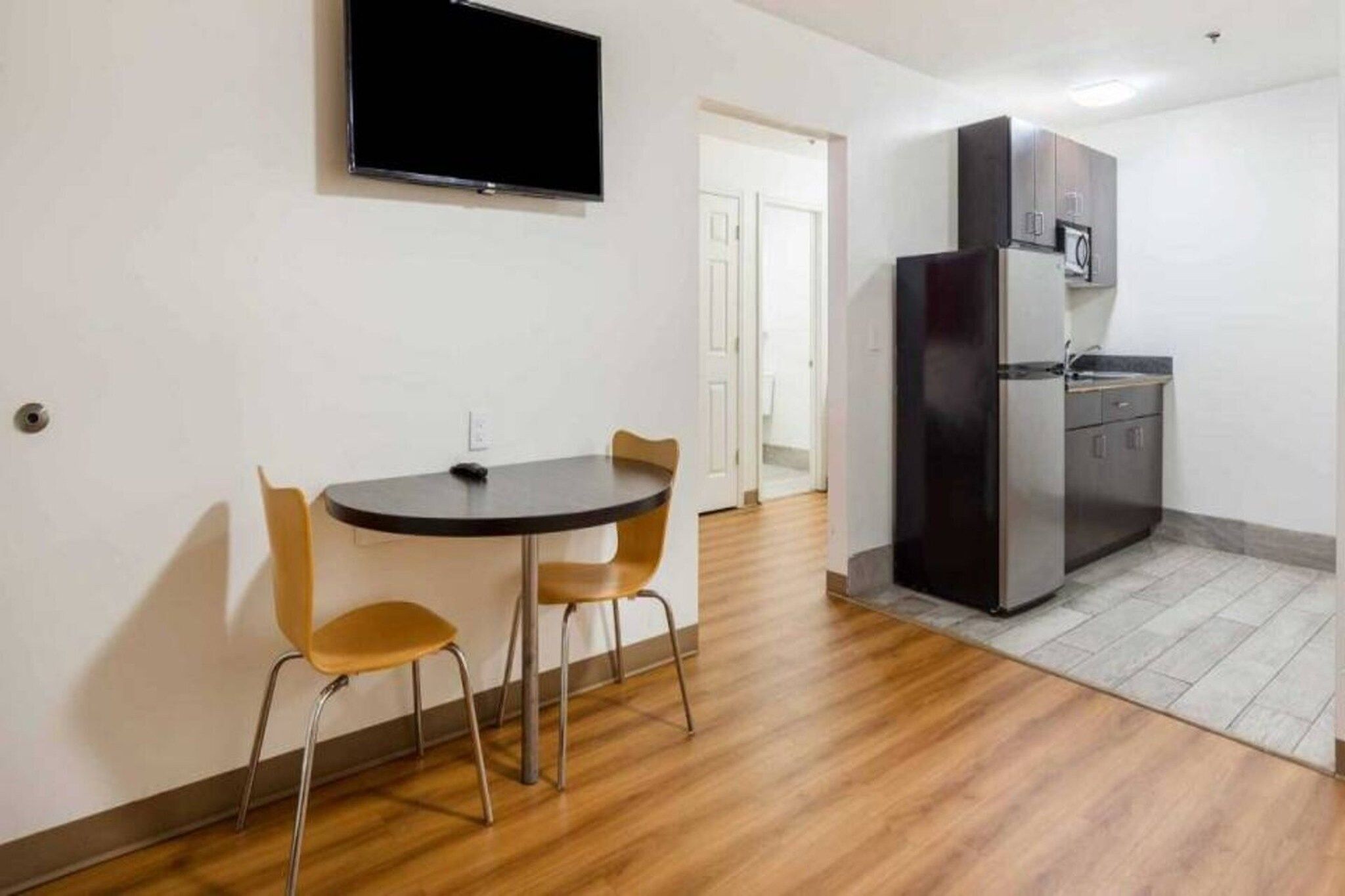 Ein kleiner Raum mit einem runden Tisch und zwei Stühlen, einer Küchenzeile inklusive Kühlschrank sowie einem an der Wand befestigten Flachbildfernseher.