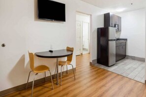 Una habitación pequeña con una mesa redonda y dos sillas, una kitchenette con refrigerador y un televisor de pantalla plana montado en la pared.