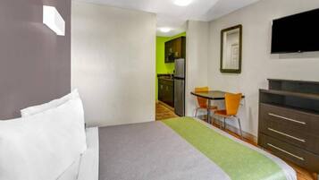 Habitación de hotel con cama, escritorio con silla, televisión y una kitchenette al fondo.