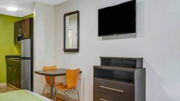 A room with a bed, a table with chairs, a television mounted on the wall, a refrigerator, and a cabinet.