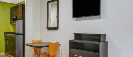 A room with a bed, a table with chairs, a television mounted on the wall, a refrigerator, and a cabinet.