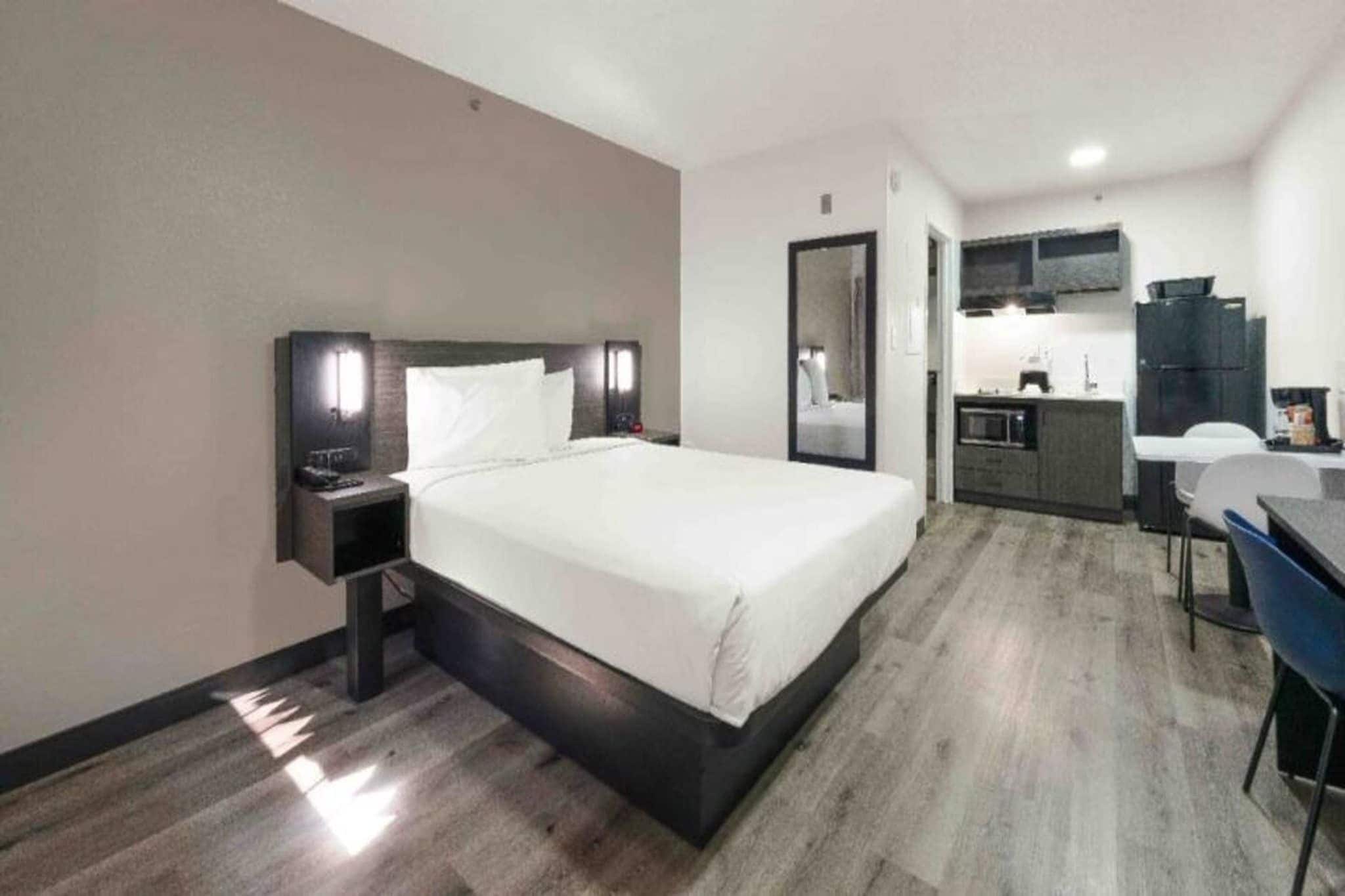 A hotel room with a bed, bedside table, a desk with a chair, a mirror, and a kitchenette area.