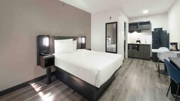 A hotel room with a bed, bedside table, a desk with a chair, a mirror, and a kitchenette area.