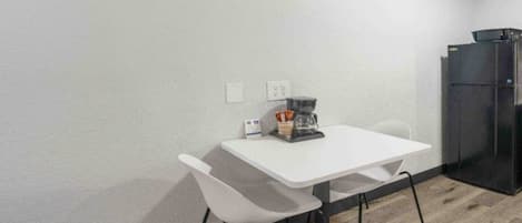 Een kleine, eenvoudige kamer met een witte tafel en twee stoelen, een koffiezetapparaat en een koelkast.