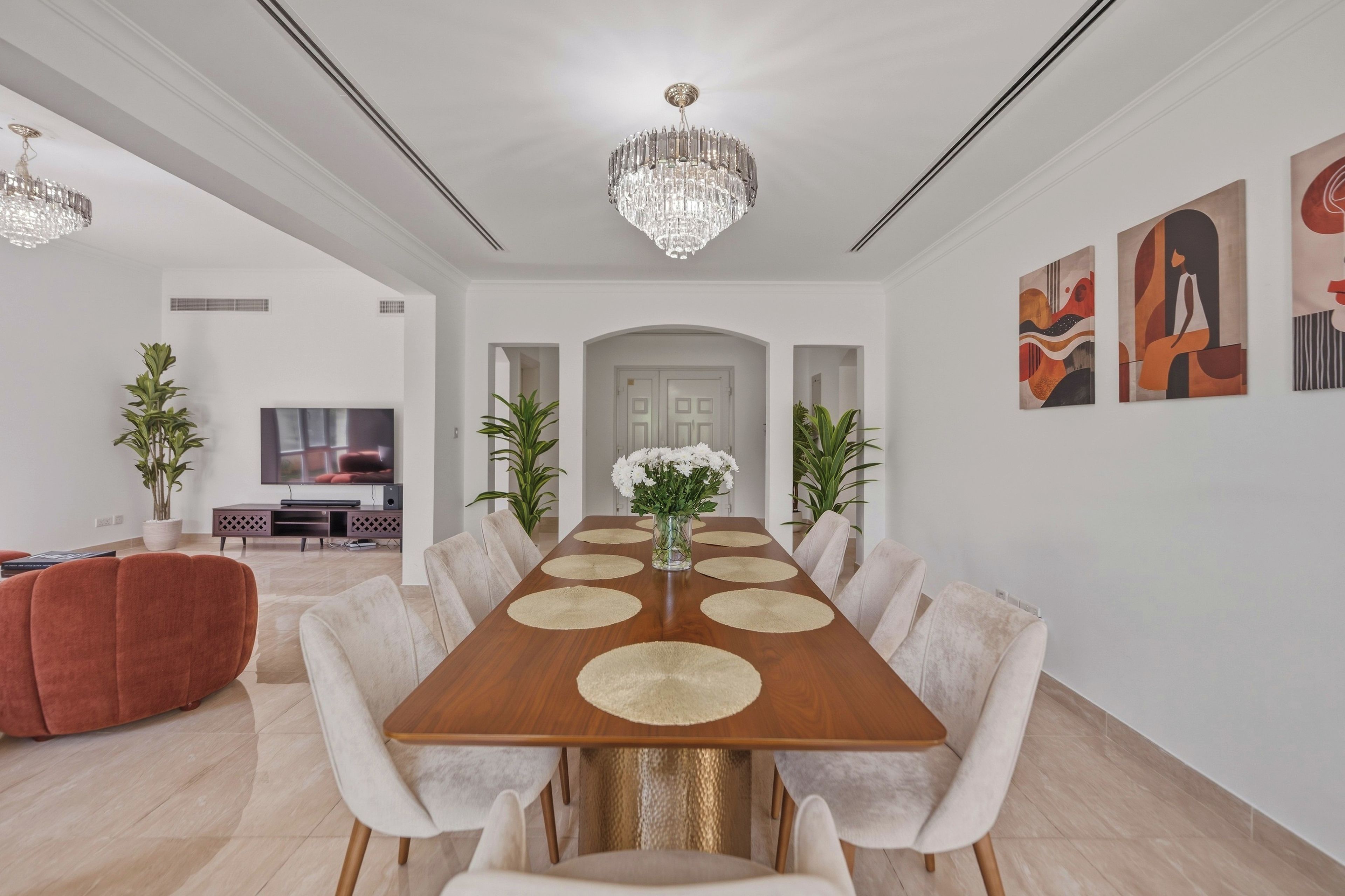 A spacious dining room with a long wooden table, white upholstered chairs, and a chandelier.