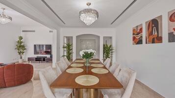 A spacious dining room with a long wooden table, white upholstered chairs, and a chandelier.