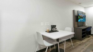 Een lichte, moderne kamer met een witte tafel, een koffiezetapparaat en een aan de muur bevestigde televisie.