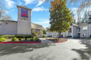 A hotel entrance with a sign for '6' and 'office' visible, surrounded by parking and greenery. - 1 Queen Bed | ADA/RI Shower (Tucker)