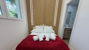 Una cama con una manta roja, almohadas blancas y toallas enrolladas.