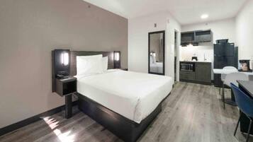 A hotel room with a bed, bedside table, a desk with a chair, a mirror, and a kitchenette area.