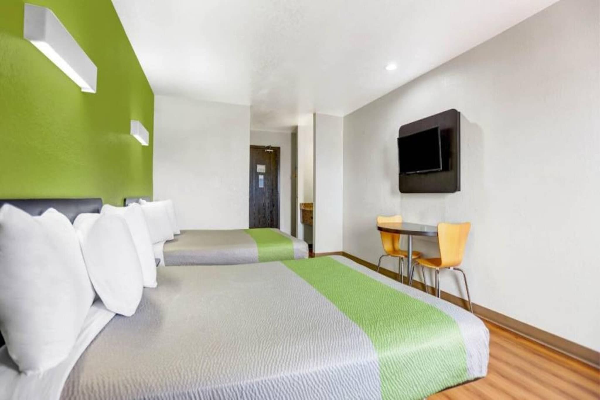A hotel room with two beds, a desk with chairs, a flat-screen TV, and a green accent wall.