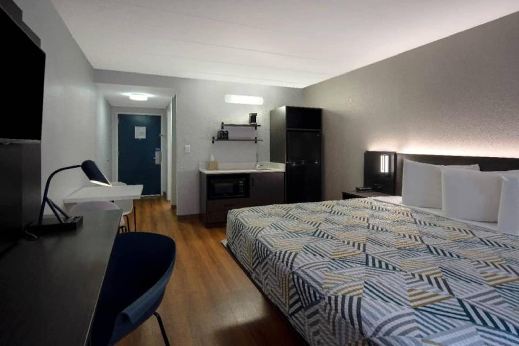 Quarto de hotel com cama, escrivaninha, cadeira, cozinha compacta e televisão.