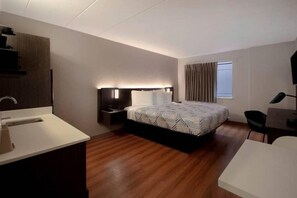 Ein modernes Hotelzimmer mit Holzboden, einem großen Bett mit gemusterten Bettbezügen, einem Nachttisch mit Lampe, einem Schreibtisch mit Stuhl und einem Fenster mit Vorhängen.