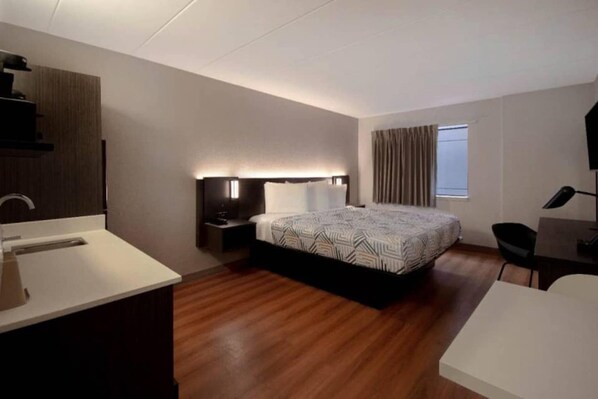 Habitación de hotel moderna con piso de madera, una cama grande con ropa de cama estampada, una mesita de noche con lámpara, un escritorio con silla y una ventana con cortinas.