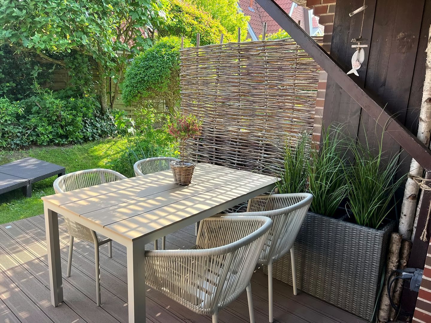 Eine Terrasse im Freien mit Tisch und Stühlen, umgeben von Pflanzen und einem Korbbodenzaun.