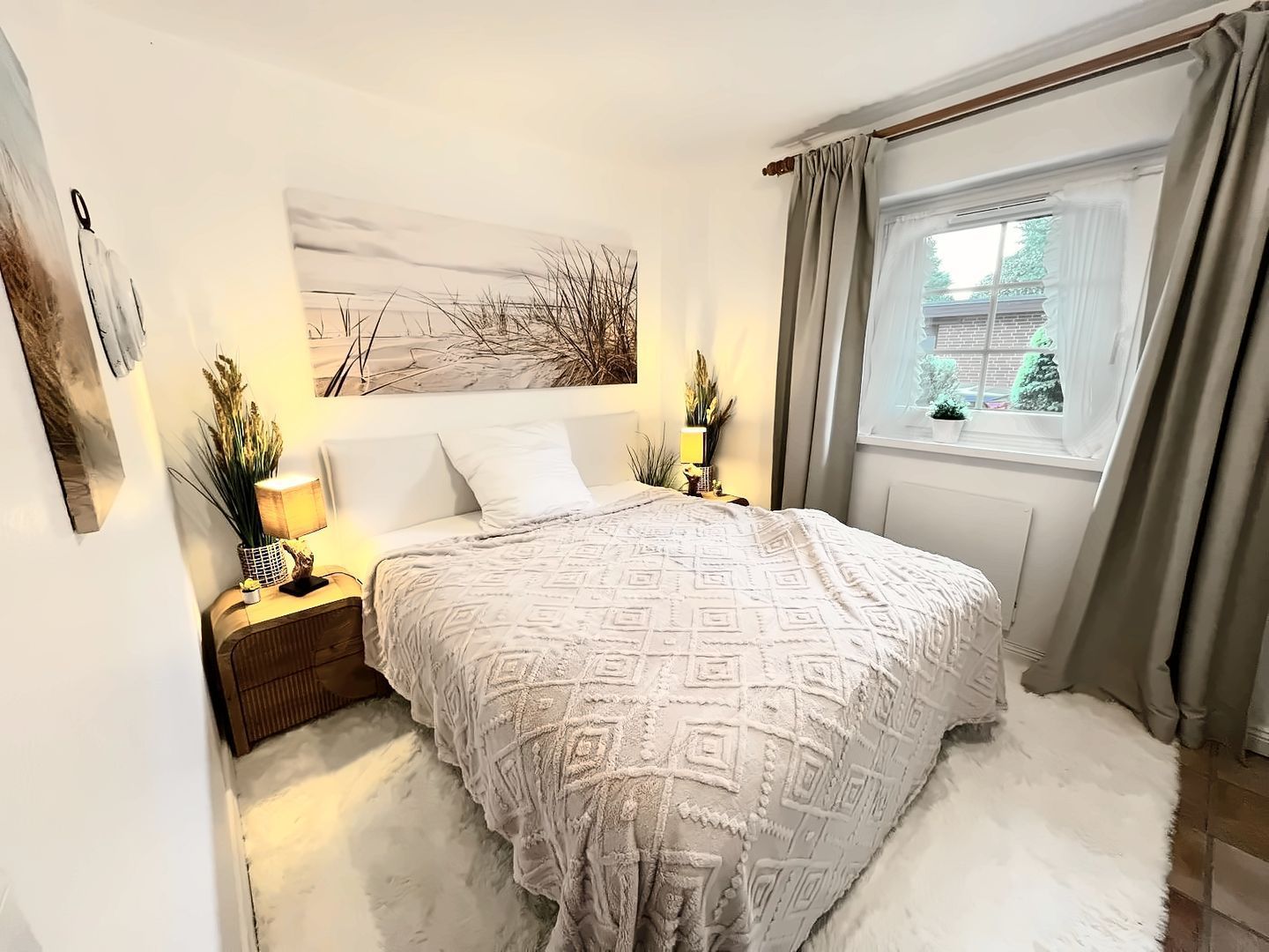 Une chambre avec un grand lit, une table de chevet, une lampe, une fenêtre avec des rideaux et un tableau au mur.