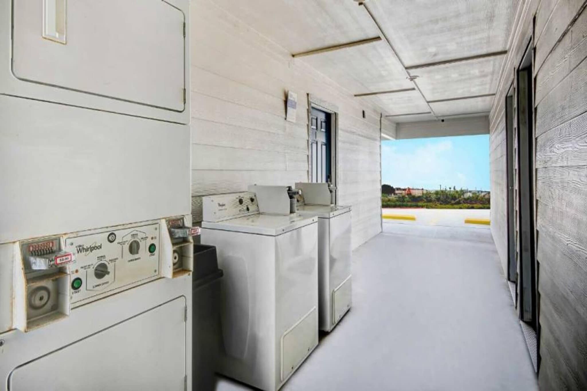 A laundry room with industrial-style washers and dryers, a control panel, and a view of the outside.