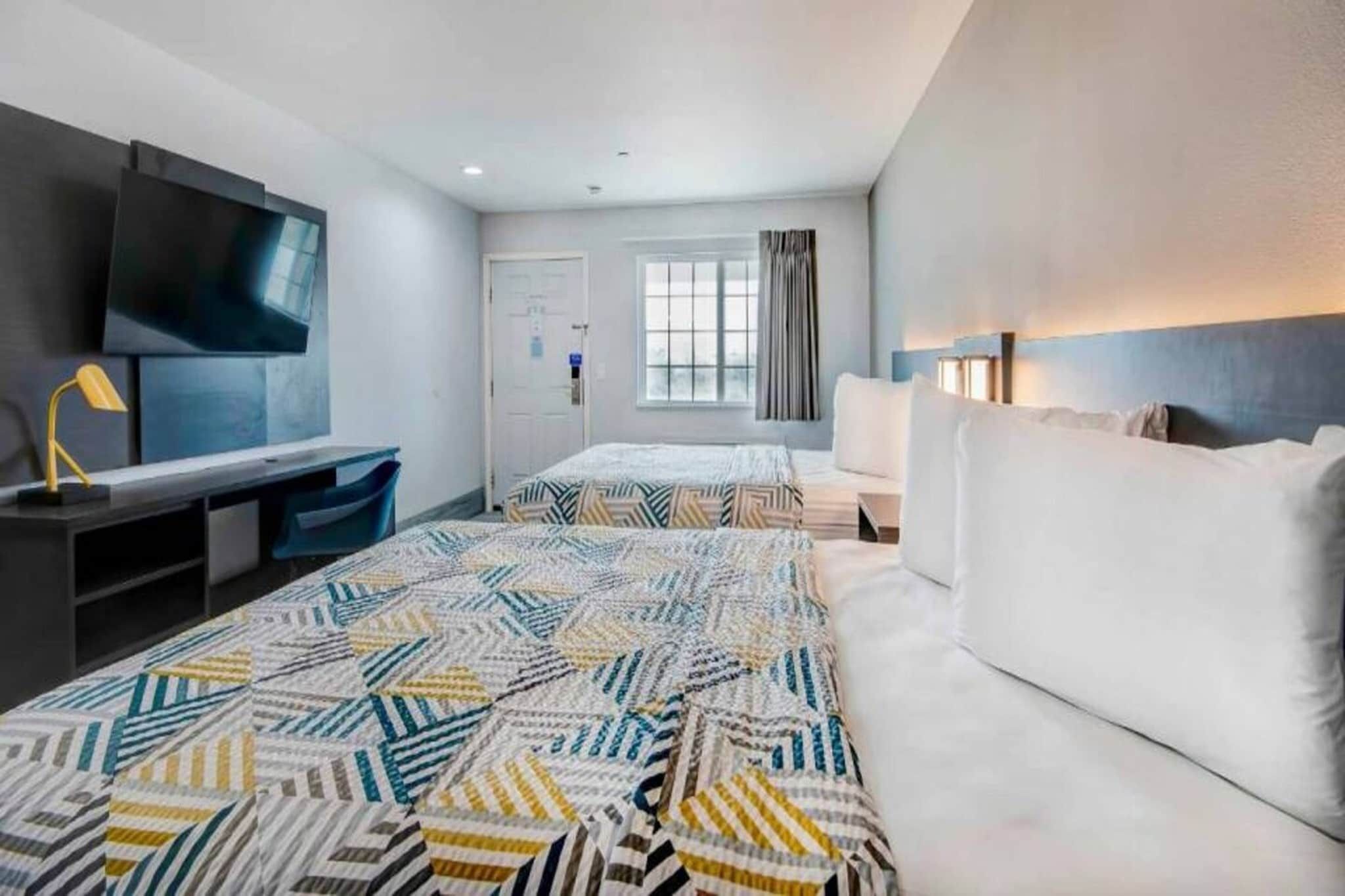 A hotel room with a large bed, a desk with a chair, a TV mounted on the wall, and a window with curtains.