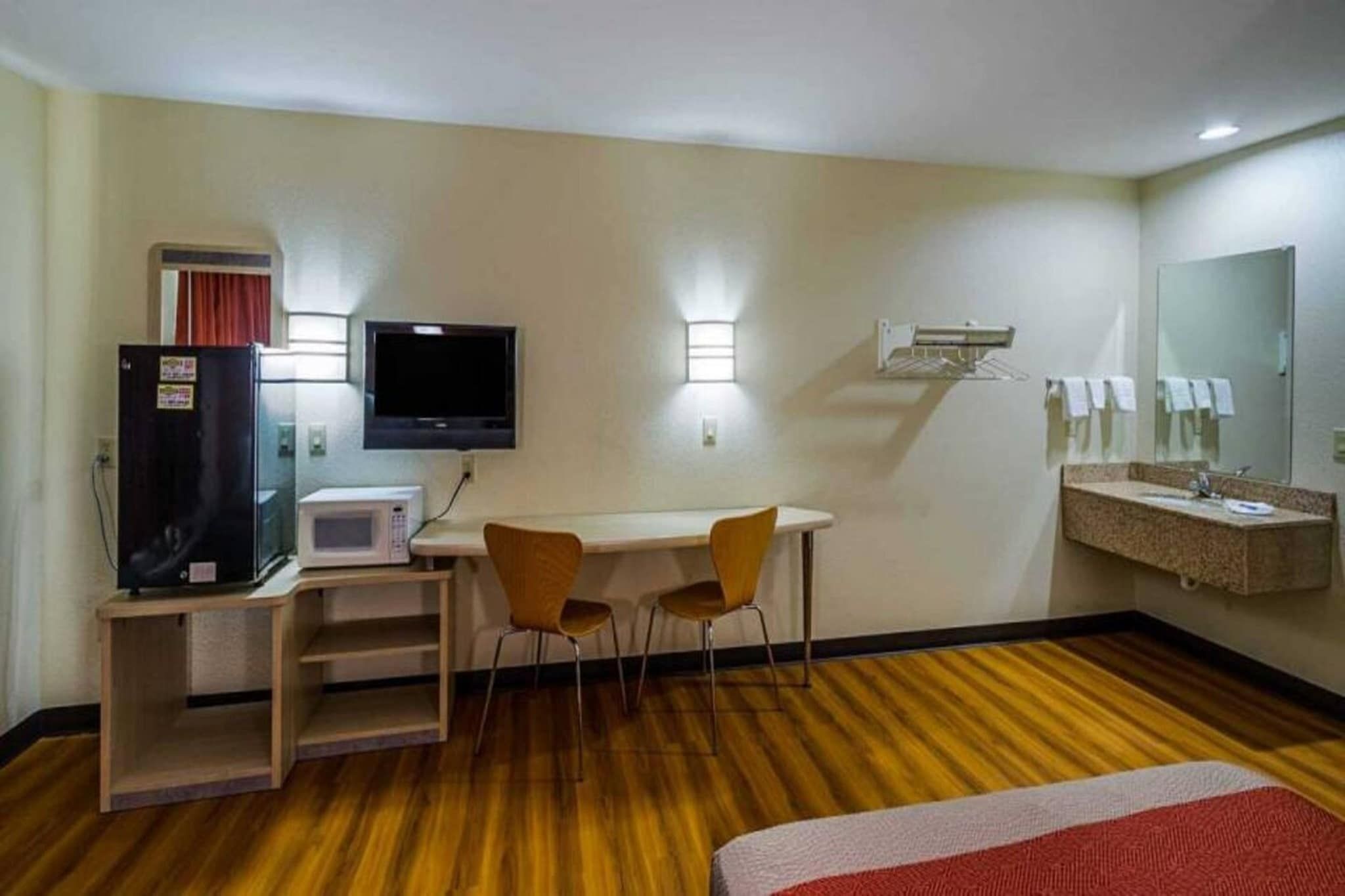 A hotel room with a bed, a desk with a computer and a TV, a microwave, a small table with chairs, a mirror, and a shower area.