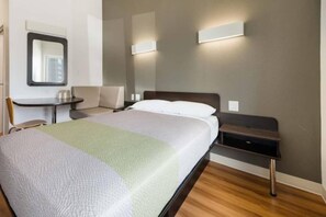 A hotel room with a bed, bedside table, chair, and a wall-mounted light fixture. - 1 Queen Bed | Mobility/Hear Access Tub Nsmk (Corpus Christi TX)