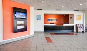 Zona de recepción de hotel con una pantalla digital, mostrador de recepción y un atril con revistas.