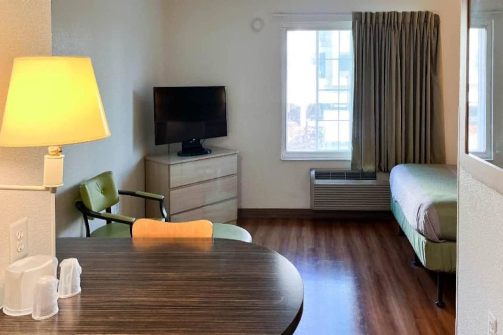 Una habitación de hotel con una mesa de comedor, un televisor, un sofá y un ventanal con cortinas.