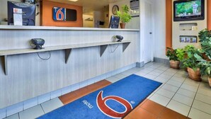 Zona de recepción de hotel con mostrador, reloj, televisor y alfombra azul con un logo.