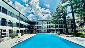 Una piscina con sillones de descanso, un edificio con varios balcones y un cielo despejado.