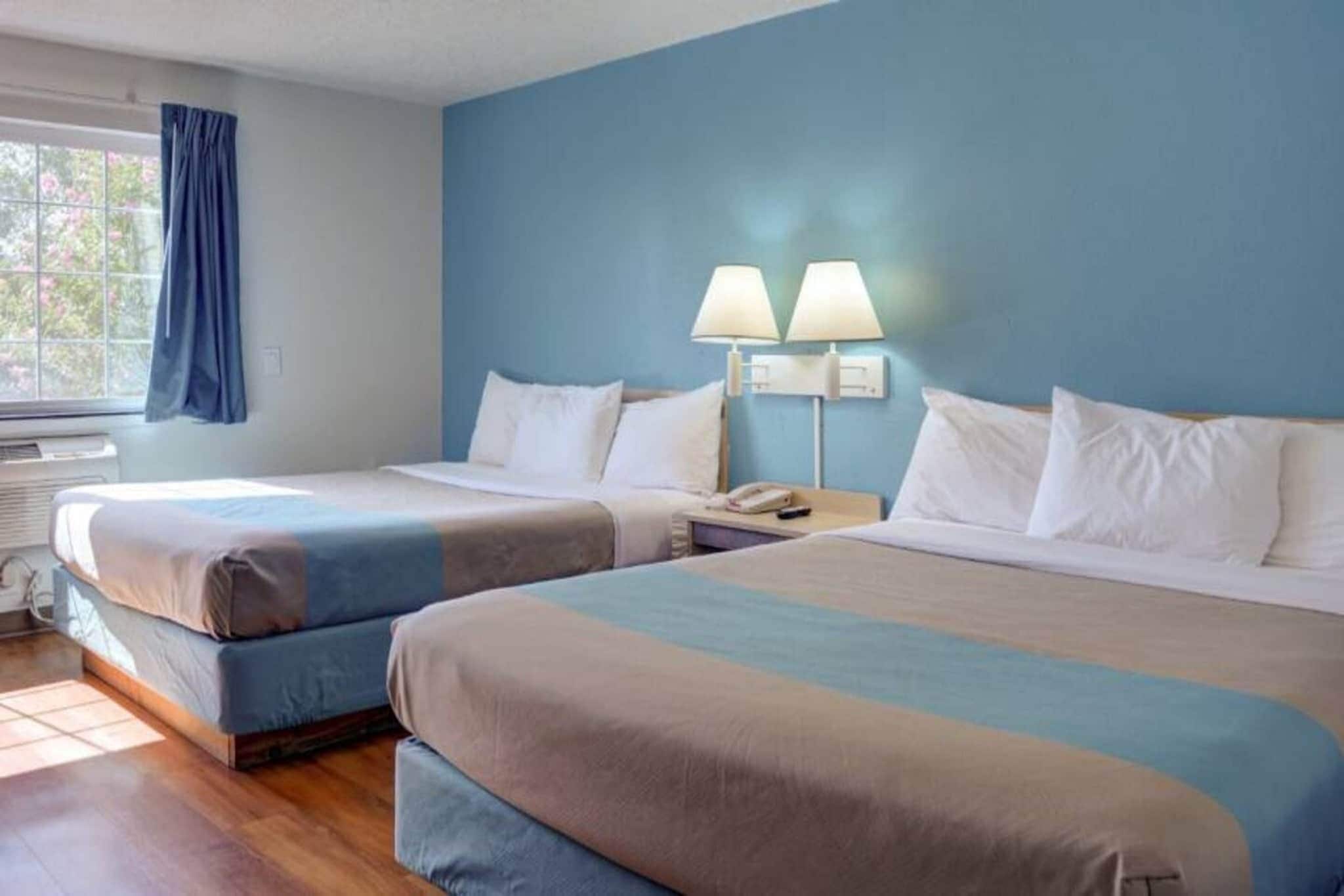 Habitació d'hotel amb dos llits, una finestra amb cortines, un aparell d'aire condicionat i una tauleta de nit amb una làmpada.