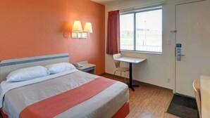 Una habitación de hotel con cama, mesita de noche, lámpara, silla, escritorio, ventana y puerta.