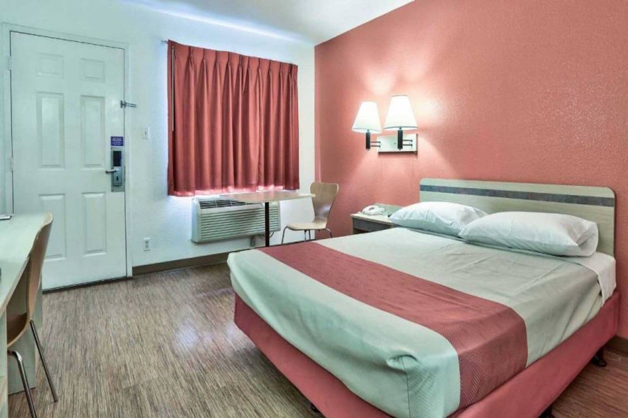 A hotel room with a bed, a desk, a chair, an air conditioning unit, and a window with curtains.