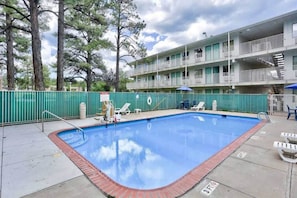 A swimming pool with a brick edge, surrounded by a green fence, and a multi-story building with balconies in the background. - 1 Queen Bed | Flagstaff AZ (Flagstaff AZ)