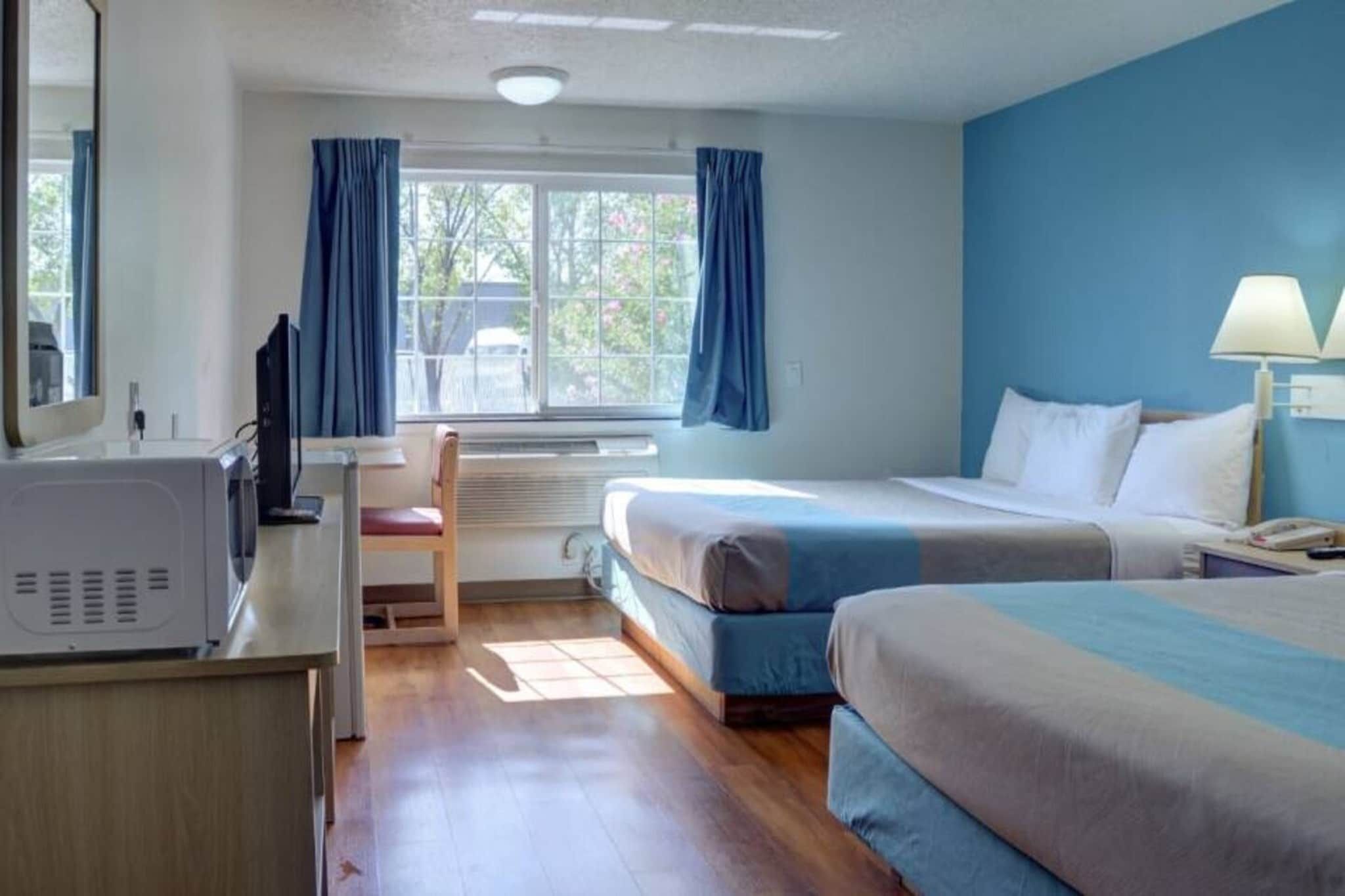 Ett hotellrum med en säng, ett skrivbord med en dator, en stol, ett fönster med gardiner och en lampa.
