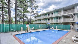 Une piscine bordée de briques, entourée d’une clôture verte, et un immeuble à plusieurs étages avec des balcons en arrière-plan.