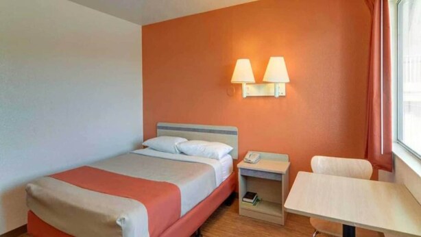 Quarto de hotel com cama, mesa de cabeceira, abajur e pequena escrivaninha com cadeira.