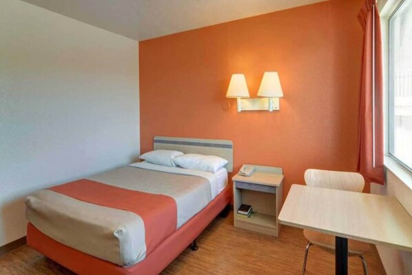 Een hotelkamer met een bed, nachtkastje, lamp en een klein bureau met stoel.
