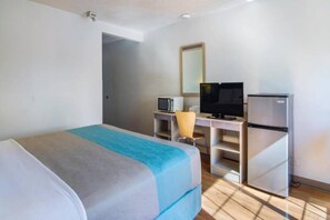 A hotel room with a bed, a desk with a computer and a television, and a refrigerator.