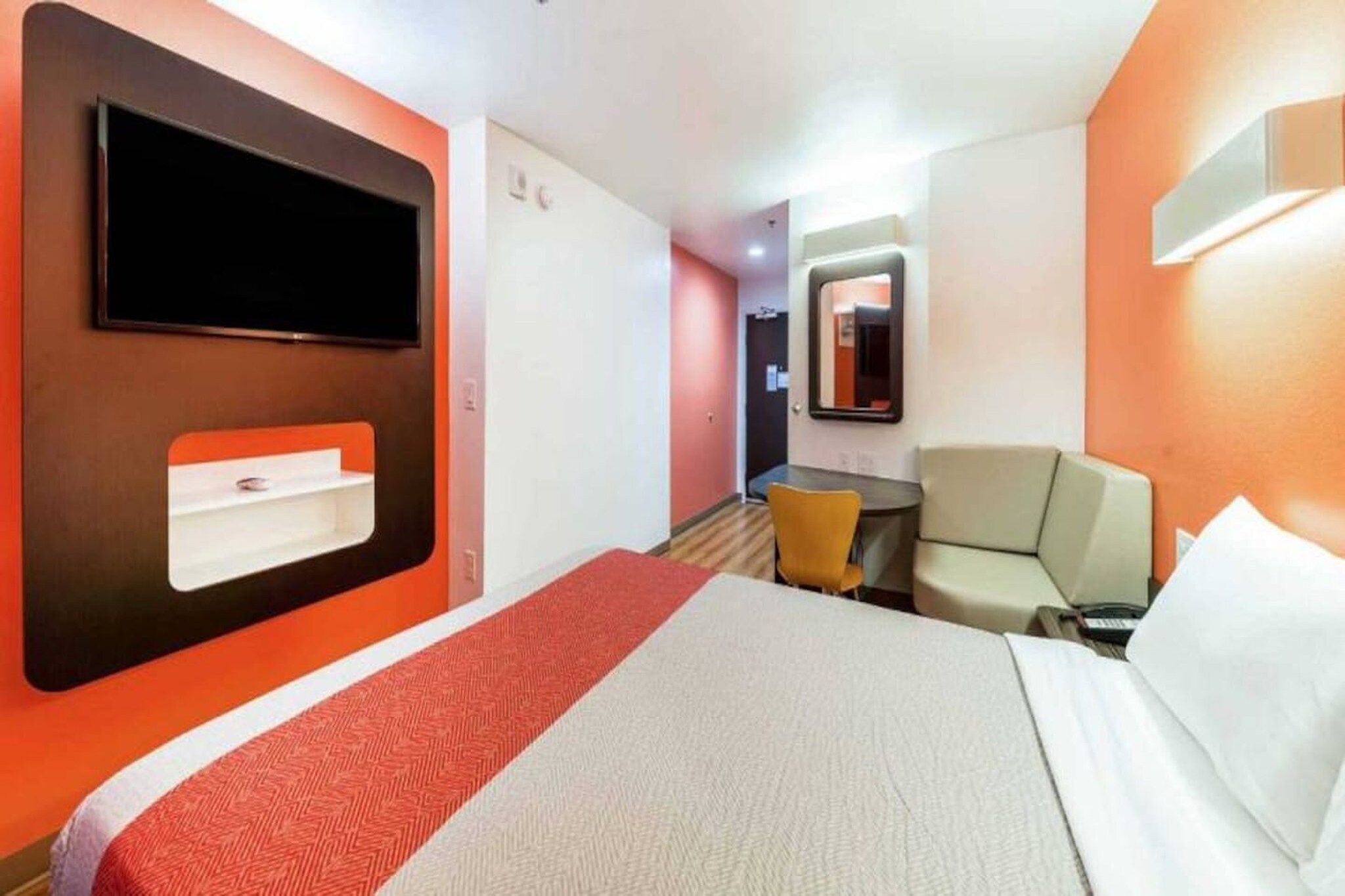 Hotellihuone, jossa on sänky, seinälle asennettu televisio, pieni pöytä ja tuoli.