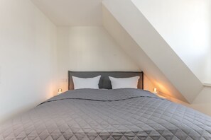 Ein Schlafzimmer mit schrägen Wänden, einem Bett mit grau gepolstertem Bettbezug, zwei Nachttischlampen und einer weißen Wand.