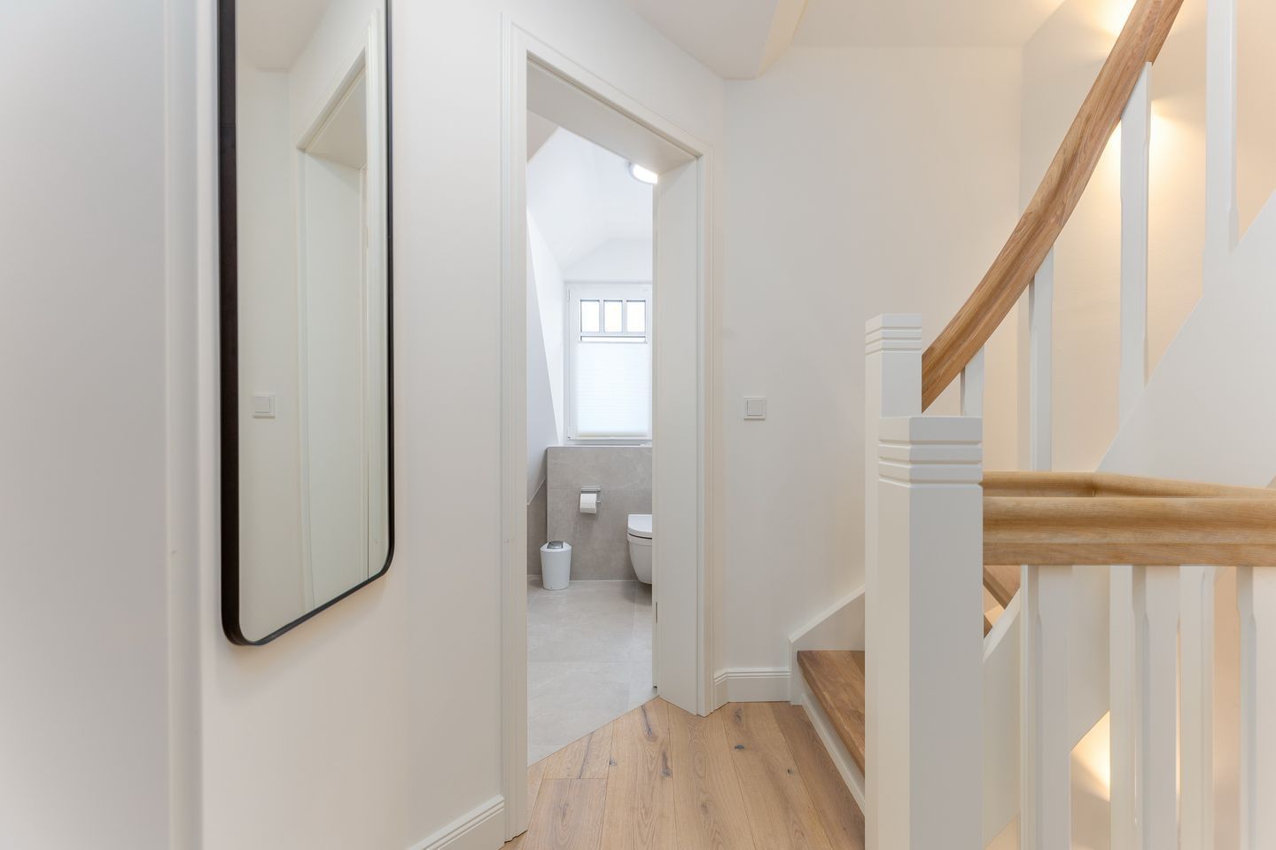 A hallway with a wooden staircase, a mirror, and a view into a bathroom with a toilet and a trash bin.