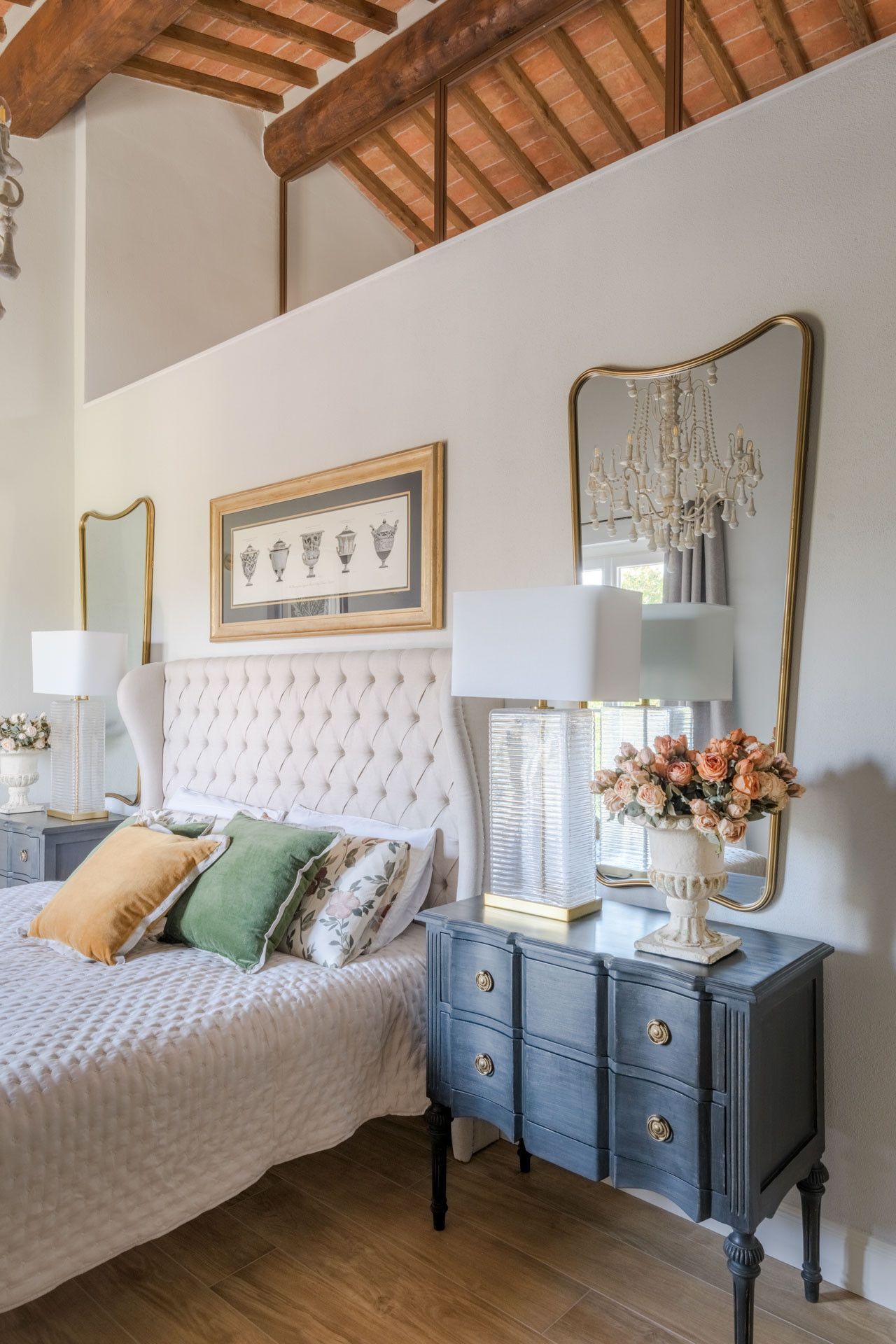 Een slaapkamer met een gestapeld hoofdeinde, een bed met een witte deken, een nachtkastje met een lamp, een dressoir met lades en een spiegel met een gouden lijst.