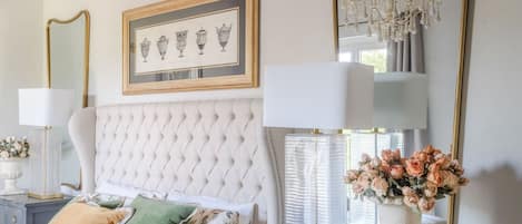 Een slaapkamer met een gestapeld hoofdeinde, een bed met een witte deken, een nachtkastje met een lamp, een dressoir met lades en een spiegel met een gouden lijst.