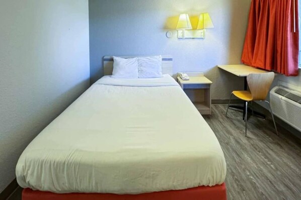Een eenpersoonsbed met een witte matras en kussens, een klein nachtkastje met een lamp, een stoel en een verwarming.