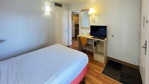 Quarto de hotel com uma cama de solteiro, uma escrivaninha com computador e uma televisão.